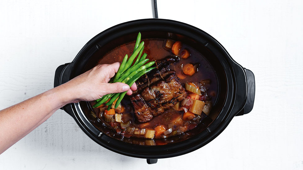 Person putting green beans into a slow cooker that has meat and vegetables inside.